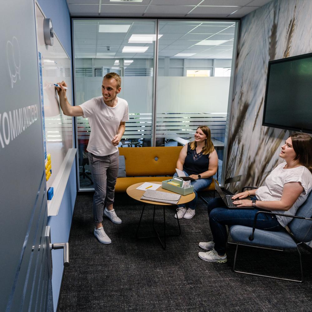 Drei Kollegen sitzen gemeinsam in einem Meetingraum mit Sofa und Sessel, einer steht am Whiteboard und schreibt.